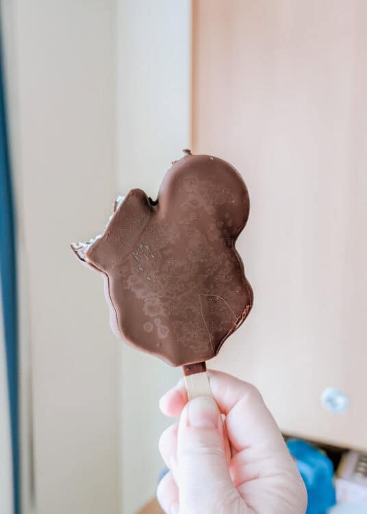 Hand holding a partially eaten Mickey Mouse-shaped ice cream bar with a chocolate coating. The iconic Disney treat is photographed indoors with a soft background of light wood and neutral walls.