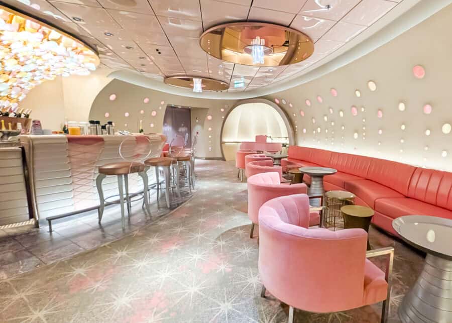 Chic and modern lounge area on a Disney cruise ship, featuring plush pink seating, a curved bar with high stools, and a wall dotted with soft-glowing circular lights. The space has a dreamy, retro-futuristic vibe with star-patterned carpet and sculptural ceiling fixtures.