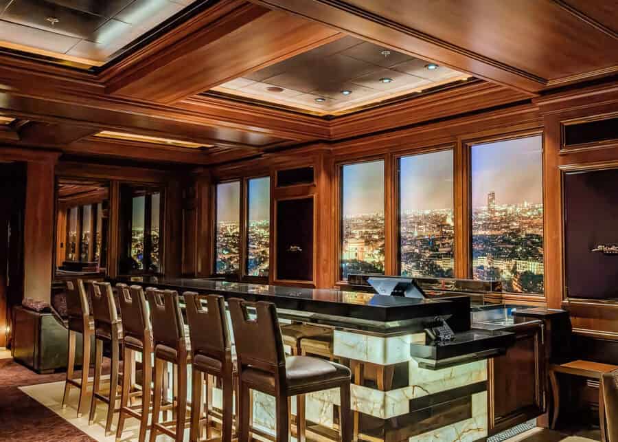 Sophisticated bar on a Disney cruise ship styled with dark wood paneling, marble accents, and high-backed leather stools, featuring a backdrop of illuminated cityscape windows depicting a night view of Paris. The moody lighting and upscale decor create an elegant, lounge-like atmosphere.