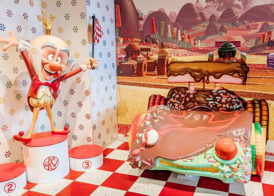 Colorful photo spot themed after Wreck-It Ralph, featuring a life-sized model of King Candy on a red and white winner’s podium next to Vanellope von Schweetz’s candy-covered race car with chocolate frosting, sprinkles, and gumdrop details. The background shows a whimsical candy racetrack scene with other racers and vibrant confectionery landscapes.