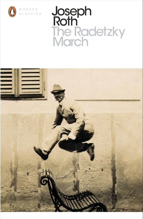 Cover of The Radetzky March by Joseph Roth, part of the Penguin Modern Classics series, featuring a sepia-toned photograph of a man in a suit and hat leaping mid-air over a curved metal bench against a stucco wall with shuttered windows. The title and author’s name appear in bold black and gray text at the top.