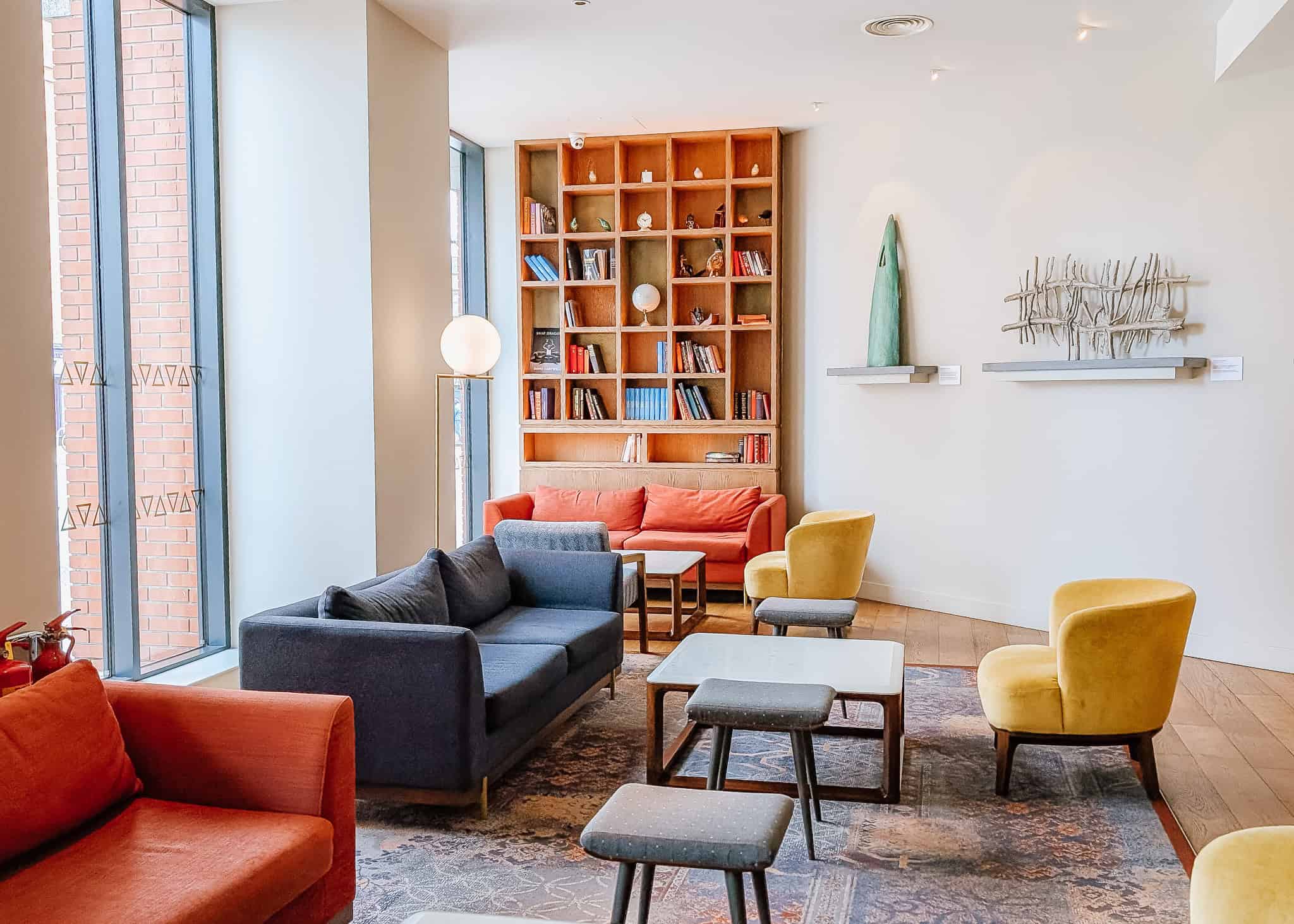 Bright and colorful hotel lounge with large windows, featuring a mix of vibrant seating including orange, navy, and mustard yellow chairs and sofas. The space includes a tall wooden bookshelf filled with books and decor, paired with minimalist wall art and soft lighting for a cozy and inviting atmosphere.