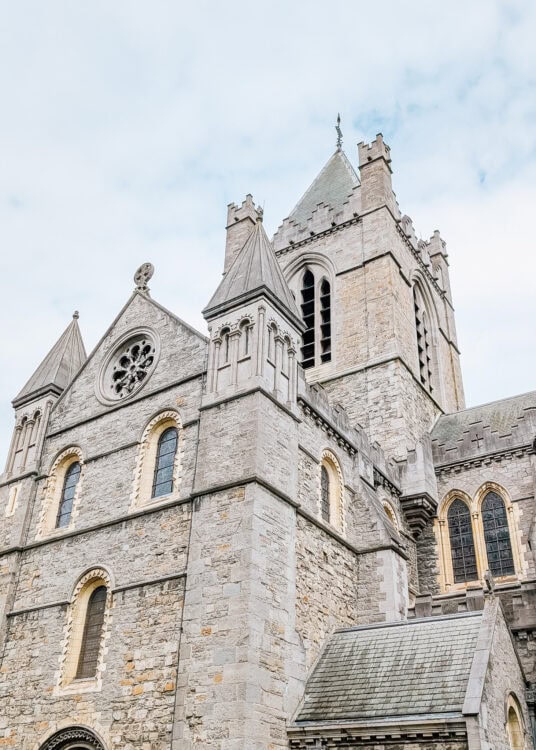 Close-up view of Christ Church Cathedral in Dublin, showcasing its gray stone exterior, pointed towers, arched windows, and Gothic architectural details. The overcast sky enhances the historic and solemn atmosphere of the medieval church.
