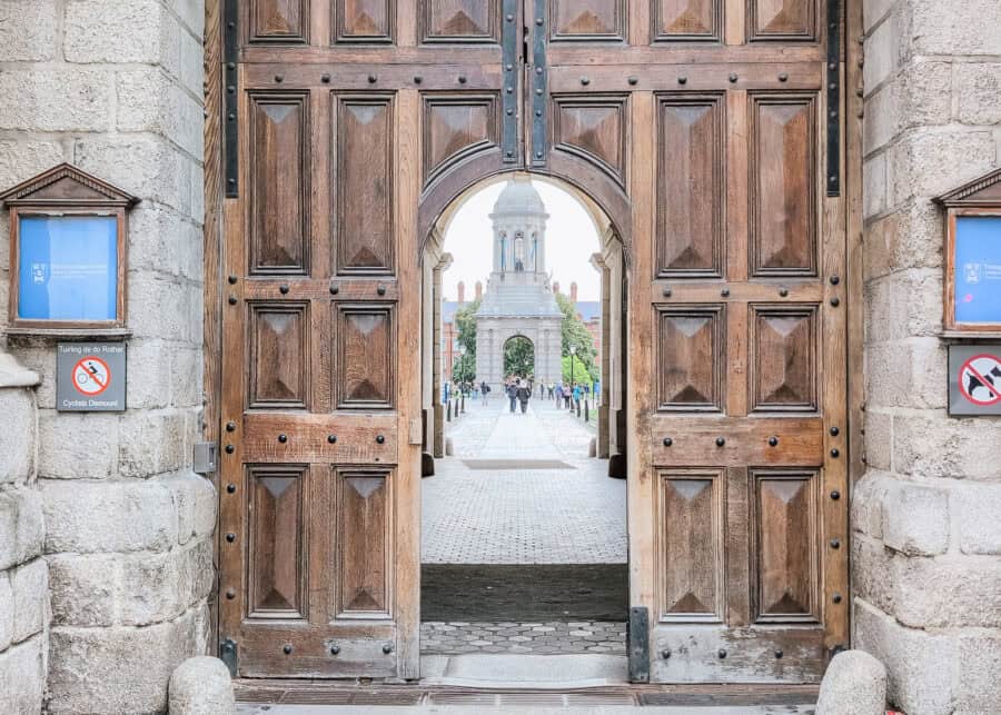 View through a large wooden gate into Trinity College Dublin, featuring the iconic Campanile bell tower framed perfectly by the arched doorway, with people walking on the cobblestone path beneath it.