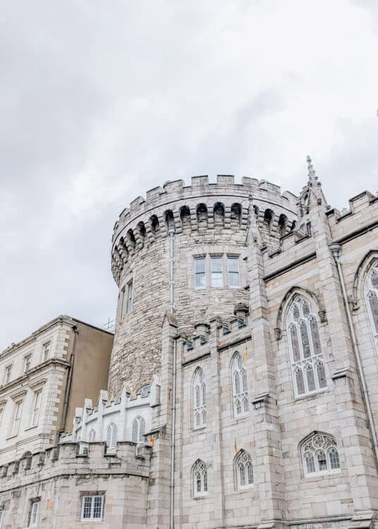 Close-up view of Dublin Castle’s round tower and adjoining Gothic Revival chapel, featuring rough stone walls, arched windows with intricate tracery, and battlement-style details. The cloudy sky adds a dramatic backdrop to the historic architecture.