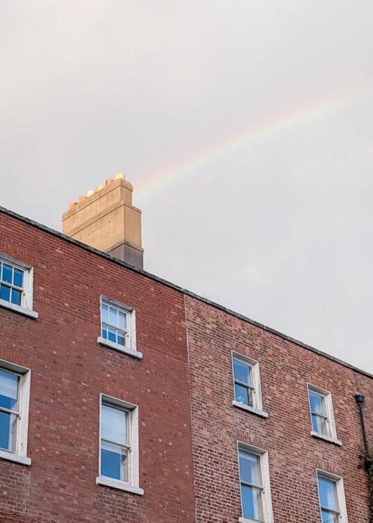 A soft rainbow arcs across a cloudy sky above a red-brick building with white-trimmed windows in Dublin. The building has a prominent chimney topped with several orange chimney pots, catching the light of the setting sun.