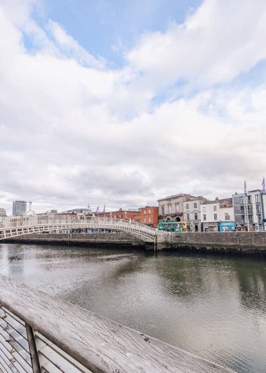 View of the Ha'penny Bridge in Dublin, an elegant white cast-iron pedestrian bridge arching over the River Liffey. Colorful buildings and a green double-decker bus line the opposite riverbank under a partly cloudy sky.