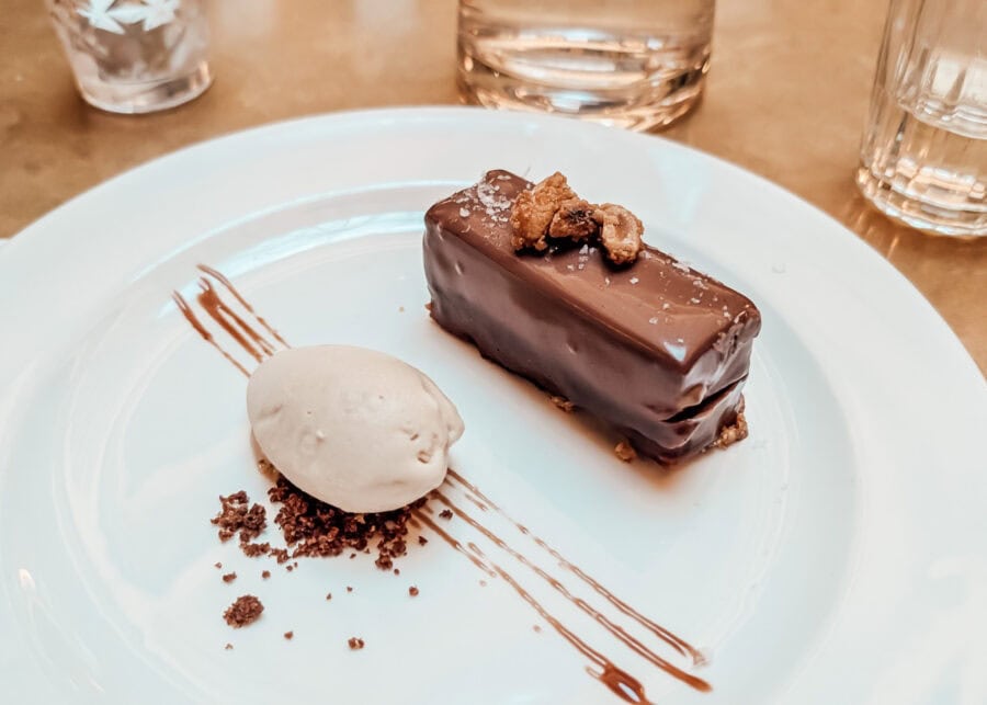 A plated dessert featuring a rectangular chocolate-covered pastry topped with nuts and a sprinkling of sea salt, served alongside a scoop of ice cream and chocolate crumbs, elegantly arranged on a white plate with chocolate drizzle.