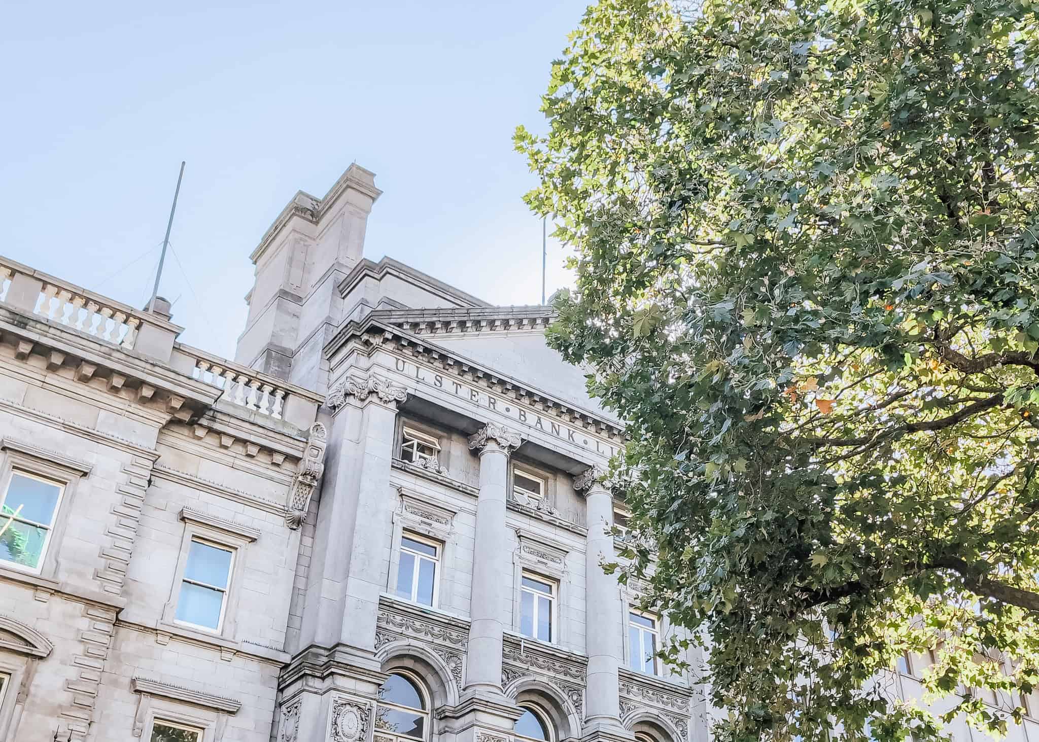 Stone‑columned neoclassical façade of an old bank building in Dublin, flanked by leafy green trees, with carved details across the lintel and “ULSTER BANK” partially visible.