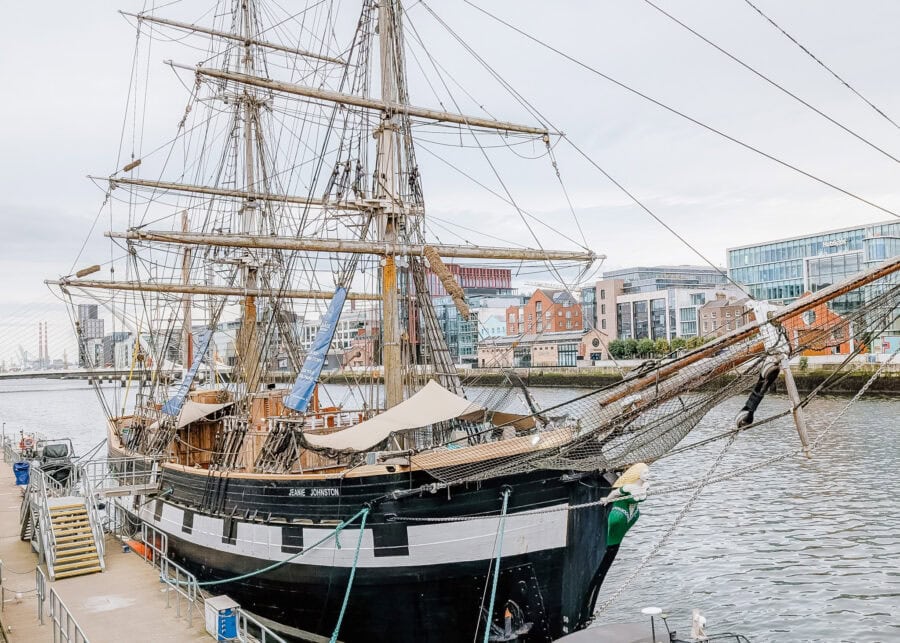 The Jeanie Johnston, a replica of a 19th-century tall ship, docked along the River Liffey in Dublin with its towering masts and intricate rigging. Modern buildings and the Samuel Beckett Bridge are visible in the background, blending maritime history with the cityscape.