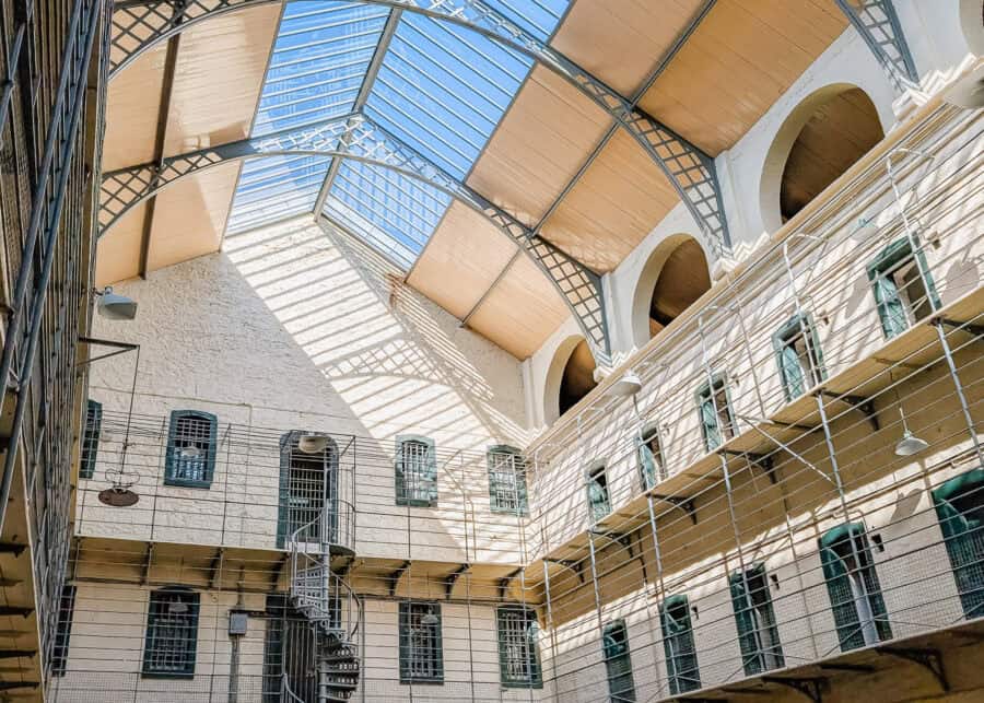 Interior view of Kilmainham Gaol in Dublin, showing a bright, open cell block with a high arched skylight ceiling casting shadows on the stone walls. Multiple rows of barred windows and green cell doors are connected by metal walkways and a spiral staircase.