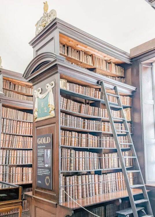 Bookshelves filled with centuries-old leather-bound books inside Marsh’s Library in Dublin. A tall ladder rests against the dark wooden shelves beneath a bright window, and a display sign titled “GOLD” honors Benjamin Guinness for his contributions to Irish heritage.