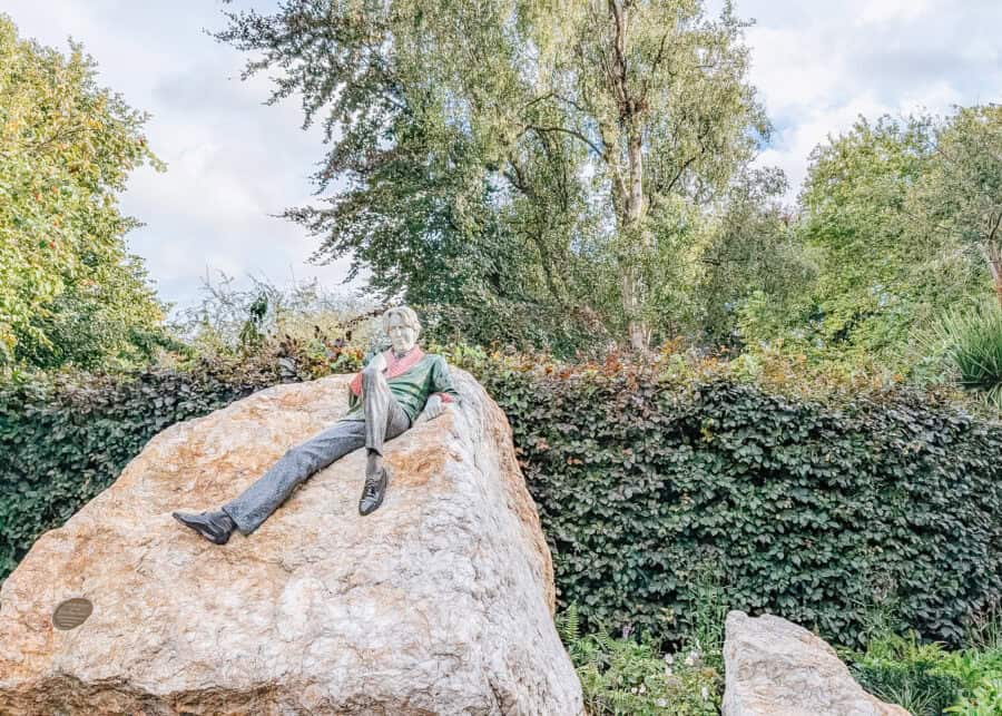 Statue of Oscar Wilde lounging on a large rock in Merrion Square Park, Dublin, with lifelike features and colorful clothing details. The sculpture is surrounded by lush greenery and ivy-covered walls, blending art with nature in a peaceful garden setting.