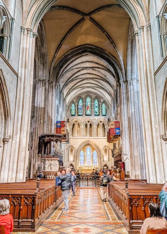 Interior of St. Patrick’s Cathedral in Dublin featuring soaring vaulted ceilings, detailed stone columns, and stained glass windows above the altar. Visitors walk along a mosaic-tiled aisle between rows of carved wooden pews, capturing the grandeur of the historic Gothic church.