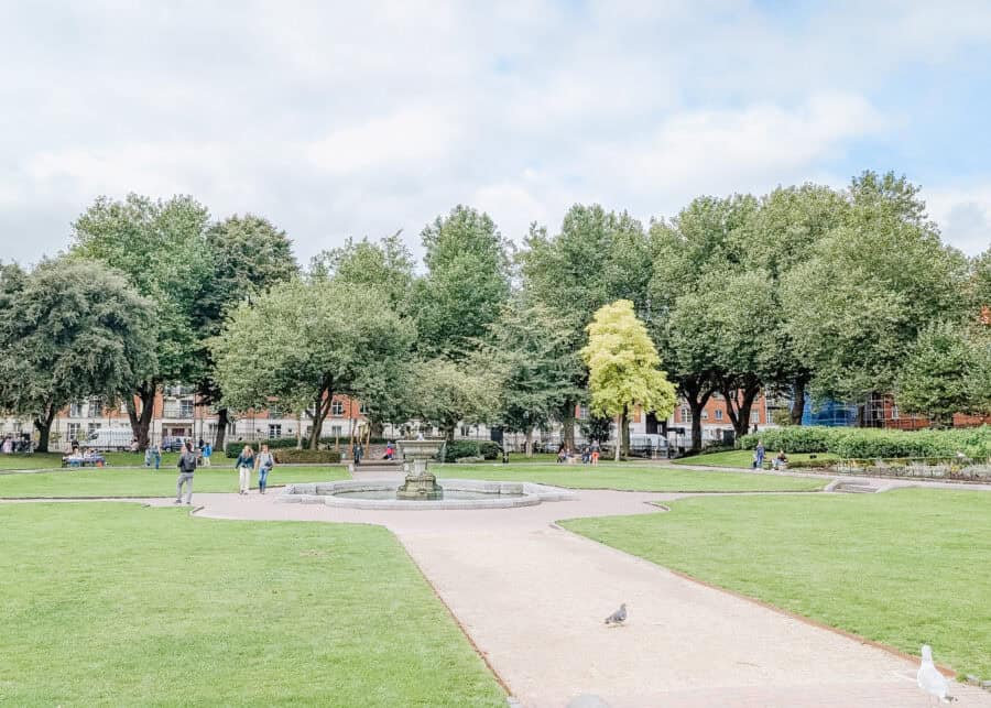 A wide view of St. Patrick’s Park in Dublin, Ireland, featuring a central stone fountain surrounded by well-kept green lawns and paved walkways. People are walking and sitting on benches, with tall leafy trees and red-brick buildings in the background. A pigeon and a seagull are visible in the foreground near the path.