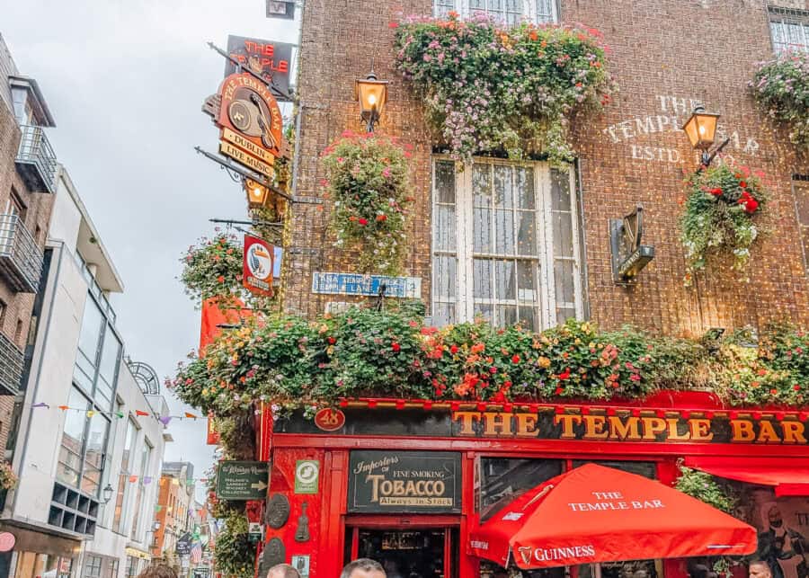 A bustling street scene at The Temple Bar in Dublin, Ireland, showing the iconic red pub adorned with vibrant flower baskets and hanging signs. The building is decorated with Guinness branding, outdoor lights, and a bright red umbrella over an outdoor seating area. The pub sits at a corner where crowds of tourists gather, and colorful flags hang between buildings down the street.