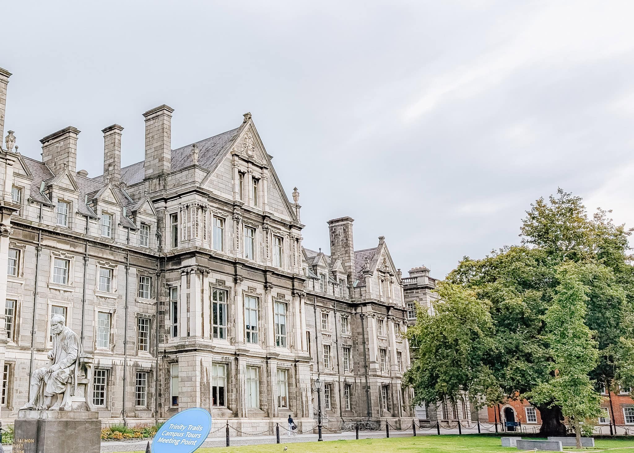 Elegant stone building on the campus of Trinity College Dublin, featuring ornate architecture with tall chimneys and decorative windows. A statue of a seated figure is visible on the lawn, near a sign reading “Trinity Trails Campus Tours Meeting Point,” with a large leafy tree adding greenery to the historic setting.