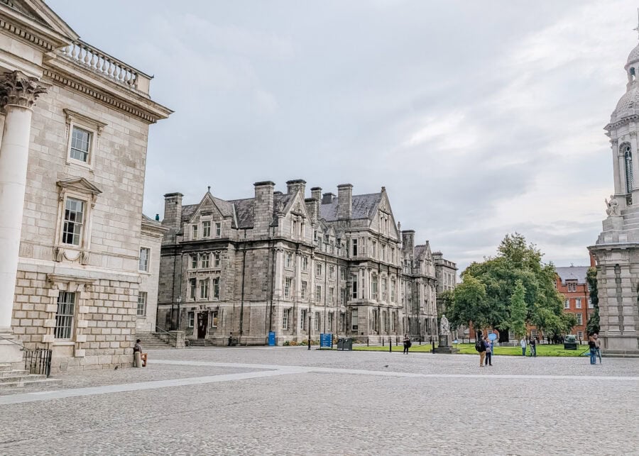 Wide view of the cobblestone courtyard at Trinity College Dublin, surrounded by grand stone buildings with classic architecture and tall chimneys. A few people stroll through the square, and statues dot the green lawn near a cluster of trees under a cloudy sky.