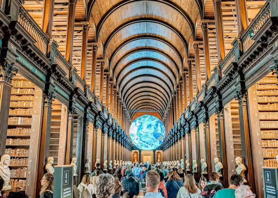 Interior view of the Long Room at Trinity College Library in Dublin, Ireland. The arched wooden ceiling stretches over tall bookshelves lined with old books and marble busts. A large illuminated globe is suspended in the center above a crowd of visitors walking through the historic space.