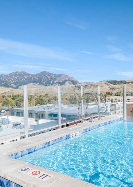 Sparkling blue rooftop pool. A glass wall encloses it, allowing swimmers to look out over the view of Bozeman mountains.