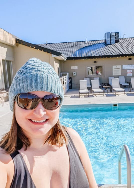 Woman wearing a knit beanie, sunglasses, and a swimsuit smiling in front of a clear blue outdoor pool on a sunny day. Lounge chairs and a building with a metal roof are visible in the background, suggesting a relaxing spa or hot spring setting.