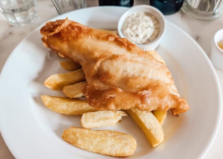 A classic serving of fish and chips on a white plate featuring a golden, crispy battered fish fillet atop thick-cut fries, with a side of creamy tartar sauce in a small ramekin.