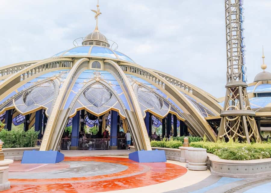 A futuristic, dome-shaped carousel attraction with a celestial and steampunk-inspired design, featuring arched golden supports, blue and gold detailing, and star motifs on the glass-like panels. The ride is partially visible through the open arches, with guests seated on stylized ride vehicles. The surrounding area includes manicured landscaping and a colorful circular plaza.