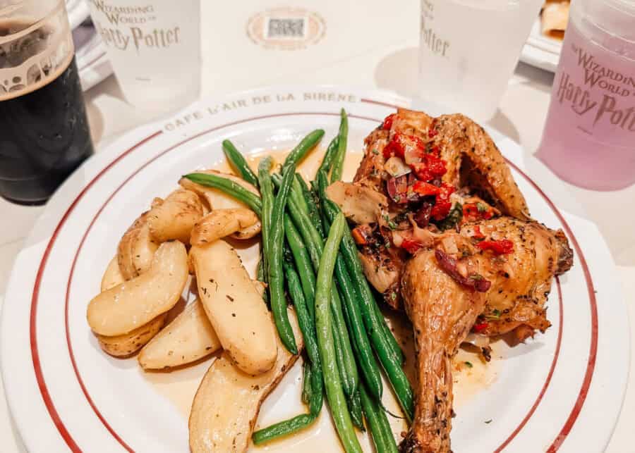 A plated meal from The Wizarding World of Harry Potter featuring herb-roasted chicken topped with a medley of sautรฉed red peppers and onions, served with green beans and roasted fingerling potatoes. The plate is set on a table with themed cups and drinks in the background, adding to the immersive Harry Potter dining experience.