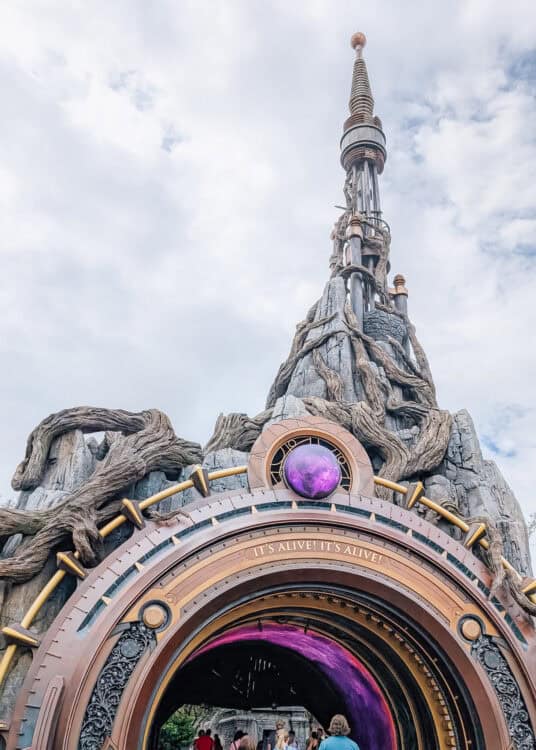A dramatic entrance to a themed attraction at Universal's Epic Universe, designed to resemble a towering, rocky structure wrapped in massive tree roots and steampunk elements. At the base is a large circular portal with a glowing purple orb and the phrase โITโS ALIVE! ITโS ALIVE!โ engraved above the archway. Visitors are walking into the illuminated tunnel beneath a cloudy sky.