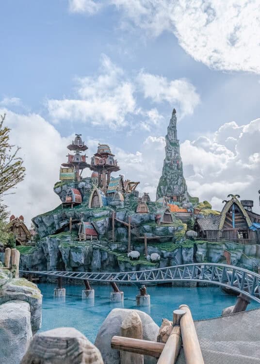 A whimsical, mountainous village scene from a theme park, inspired by Viking or fantasy architecture, featuring colorful, thatched-roof buildings perched on rocky cliffs. A tall, jagged peak rises in the background, while a bright blue waterway and a roller coaster track run in the foreground. The setting is vibrant and immersive, resembling a setting from โHow to Train Your Dragon.โ