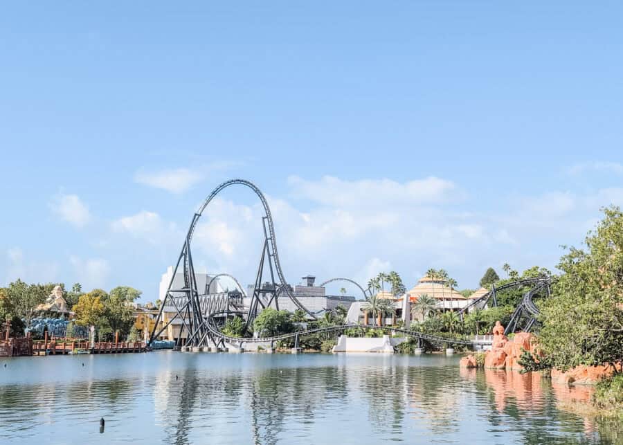 Sweeping view of the Jurassic World VelociCoaster at Universal’s Islands of Adventure, with its towering black track looping over a serene lagoon. The sleek roller coaster twists through lush palm trees and past the iconic Jurassic Park Discovery Center under a clear blue sky.