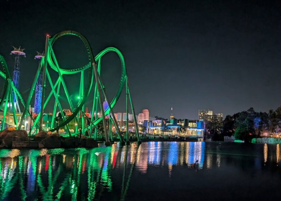 The Incredible Hulk Coaster glows bright green at night at Universal Orlando, its looping track towering over the water. CityWalk buildings and lights shine in the background, reflecting in the calm lagoon below.