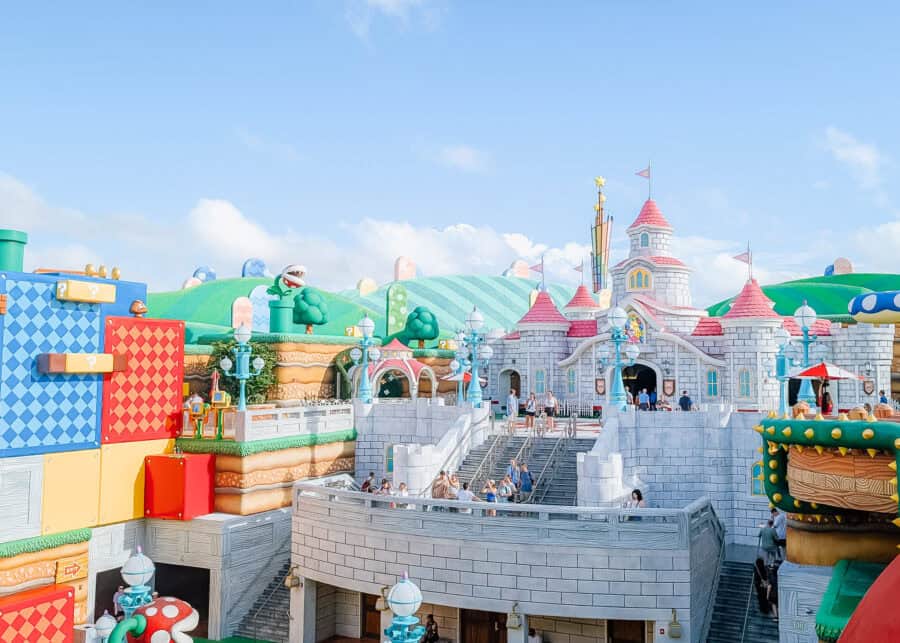 Vibrant Super Nintendo World area at Universal Studios featuring Princess Peachโs pink-roofed castle surrounded by colorful hills, giant mushrooms, and game-like elements such as question blocks and Piranha Plants. Guests walk along multi-level platforms styled like a real-life video game world under a bright blue sky.