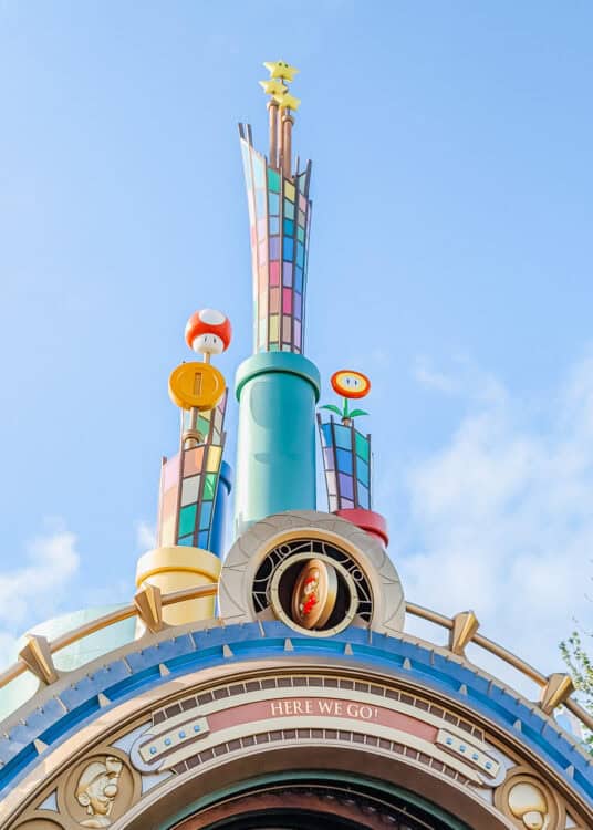 A colorful, whimsical entrance structure inspired by the Super Mario franchise, featuring bright pipes, a rainbow-tiled tower, and iconic Mario elements like a red mushroom, fire flower, gold coin, and stars at the top. The archway below includes the phrase โHERE WE GO!โ above a rounded design that resembles a portal, with playful Mario-themed details throughout.