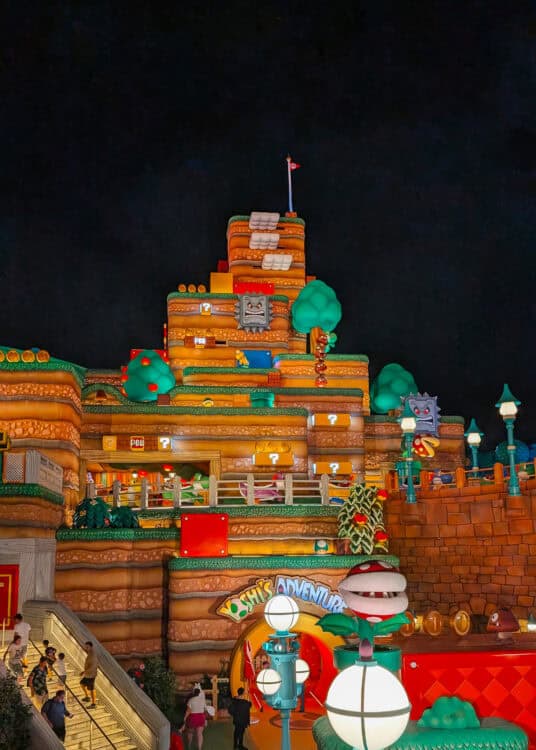A vibrant nighttime view of the Super Nintendo World attraction, themed after the Mario video game series. The scene features a multi-level, colorful mountain structure with iconic elements like ? blocks, coins, POW blocks, and characters including Goombas, Piranha Plants, and Thwomps. At the base is the entrance to โYoshiโs Adventure,โ lit with bright signage and whimsical lighting, creating a playful and immersive environment.