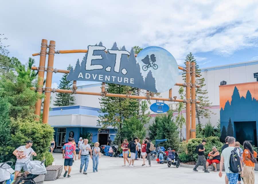Entrance to the E.T. Adventure ride at Universal Studios with a large wooden sign reading โE.T. Adventureโ above a walkway. Families with strollers and children gather beneath the sign, highlighting a popular ride for kids at Universal Studios.