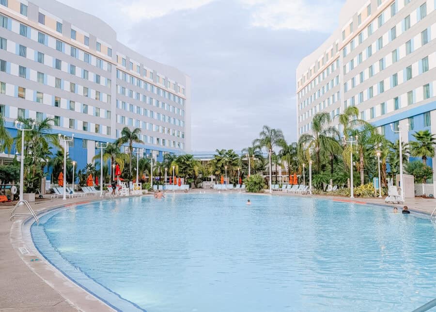 A large resort-style pool sits between two tall hotel buildings at Universal Orlando, surrounded by palm trees, lounge chairs, and bright orange umbrellas with guests swimming and relaxing.
