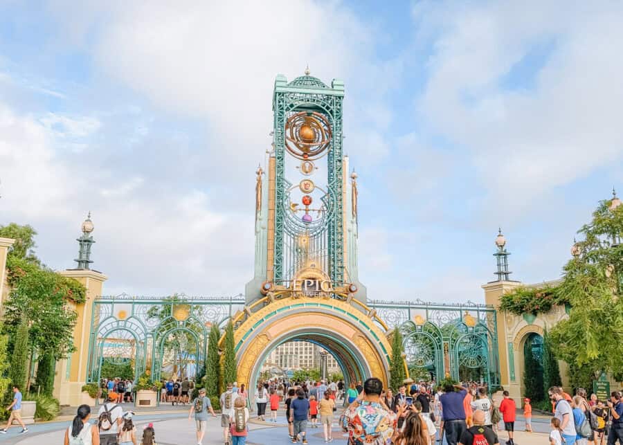 Crowds of visitors walk toward the grand entrance portal of Universalโs Epic Universe, featuring an ornate teal and gold arch with celestial designs and a tall decorative tower rising above the gateway under a bright blue sky.