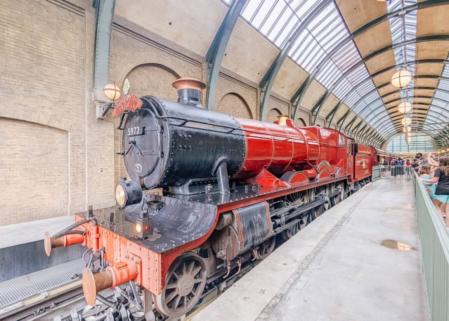 Bright red Hogwarts Express steam train parked at a station platform inside Universal Studios with guests lined up along the side. The trainโs detailed engine and carriages sit under a high arched glass ceiling, showing a popular attraction area for families and kids.