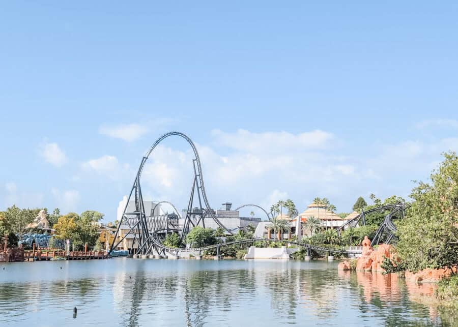 Sweeping view of the Jurassic World VelociCoaster at Universalโs Islands of Adventure, with its towering black track looping over a serene lagoon. The sleek roller coaster twists through lush palm trees and past the iconic Jurassic Park Discovery Center under a clear blue sky.