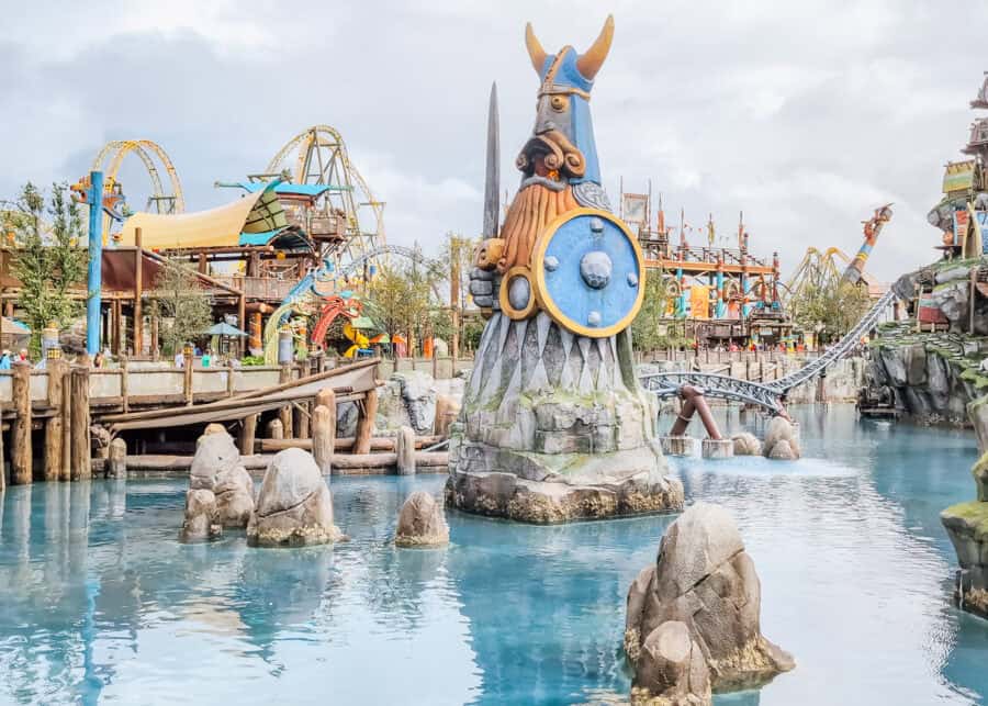 A large Viking statue holding a sword and shield rises from the water in the Isle of Berk area at Universalโs Epic Universe, surrounded by wooden walkways, themed buildings, and roller coaster tracks in the background.