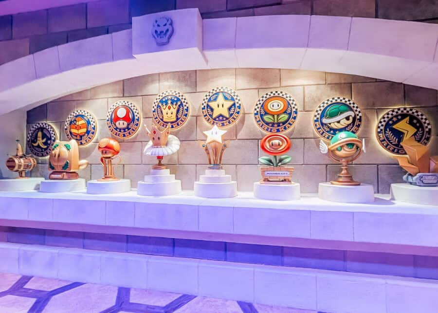 Display of Mario Kart themed trophies and power up icons including mushrooms, stars, and shells arranged on pedestals inside a Universal Studios attraction queue. Colorful circular emblems line the wall behind the figures, creating a playful indoor scene that appeals to kids and gaming fans.