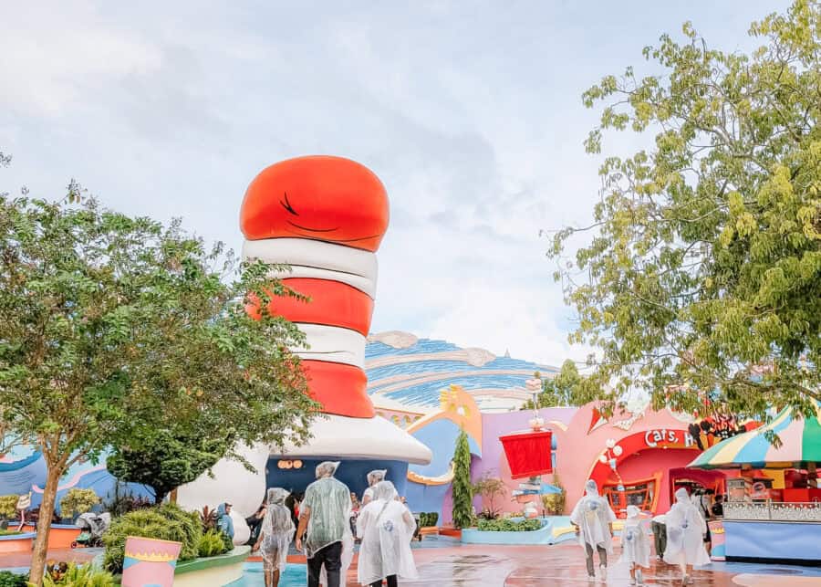Guests wearing rain ponchos walk through the colorful Seuss Landing area at Universalโs Islands of Adventure, passing the oversized red-and-white Cat in the Hat hat building and whimsical Dr. Seussโinspired architecture.