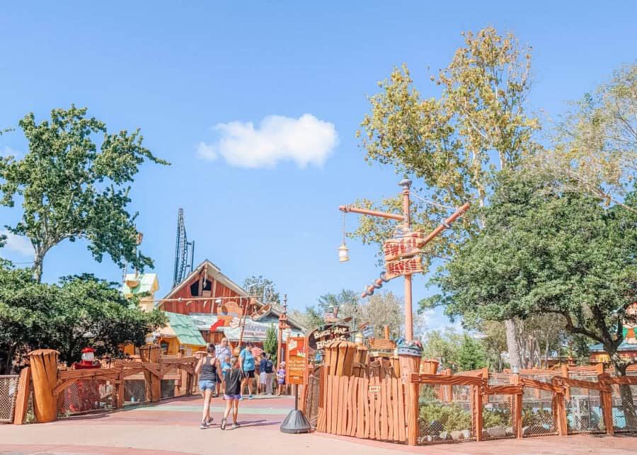 Sunny view of Toon Lagoonโs cartoon-style lumber camp area at Universalโs Islands of Adventure, featuring the entrance to Me Ship, The Olive and Popeye & Blutoโs Bilge-Rat Barges. The playful, hand-drawn aesthetic includes wooden signs, colorful buildings, and whimsical props inspired by classic comic strip characters.