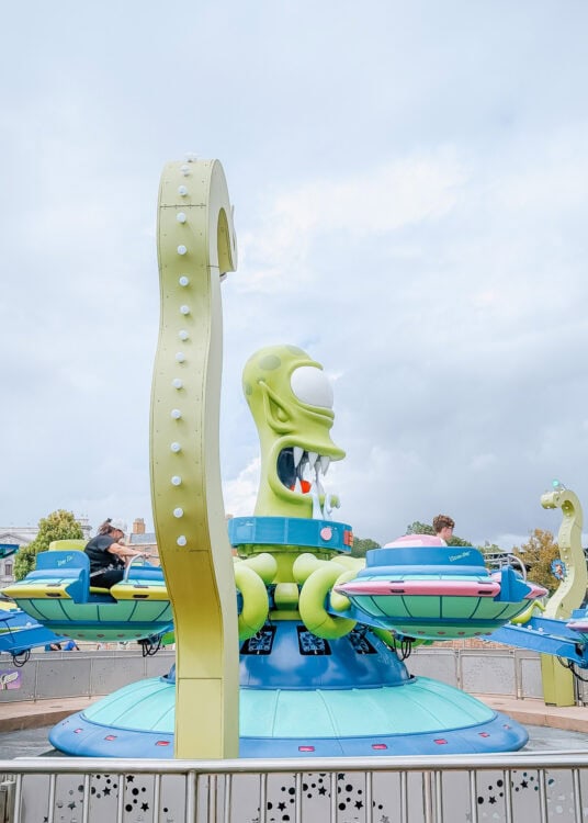 A colorful alien themed spinning ride features a large green creature with tentacles in the center while guests sit in small circular cars around it, creating a playful and whimsical amusement park scene.