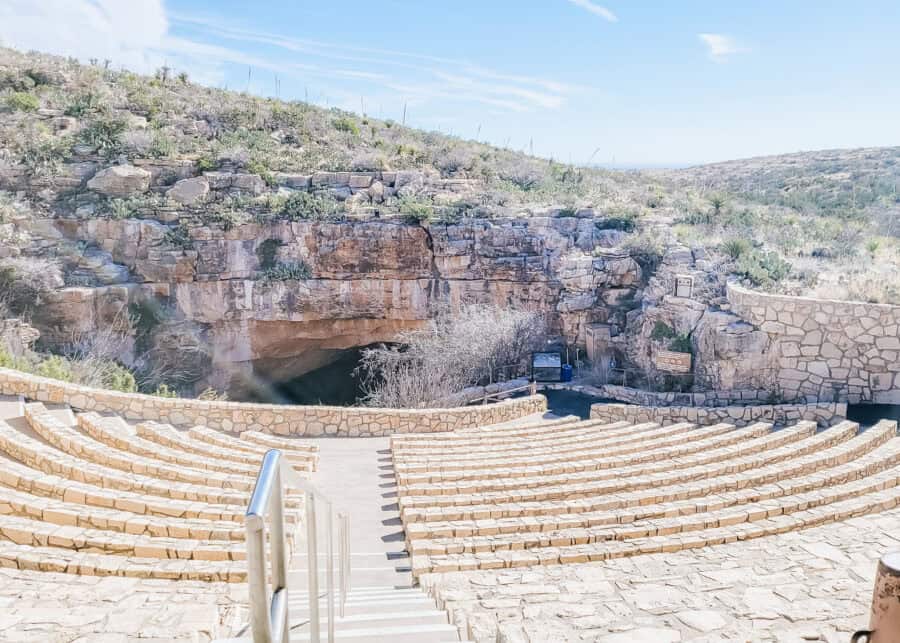 Stone amphitheater seating overlooks the natural cave entrance at Carlsbad Caverns, surrounded by desert vegetation and layered rock cliffs. The viewpoint highlights where visitors gather as part of a Carlsbad Caverns itinerary to watch bats emerge at sunset.
