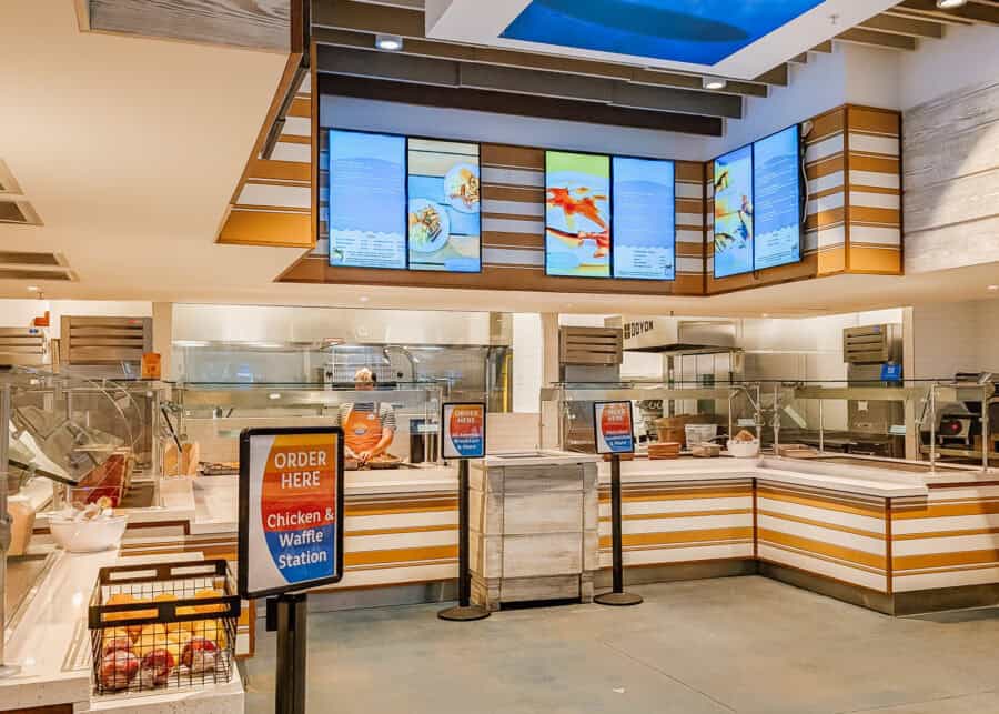 Casual cafeteria style dining area with a counter service kitchen and digital menu boards displaying food options above. A sign in front reads "ORDER HERE Chicken & Waffle Station" while staff prepare meals behind the counter