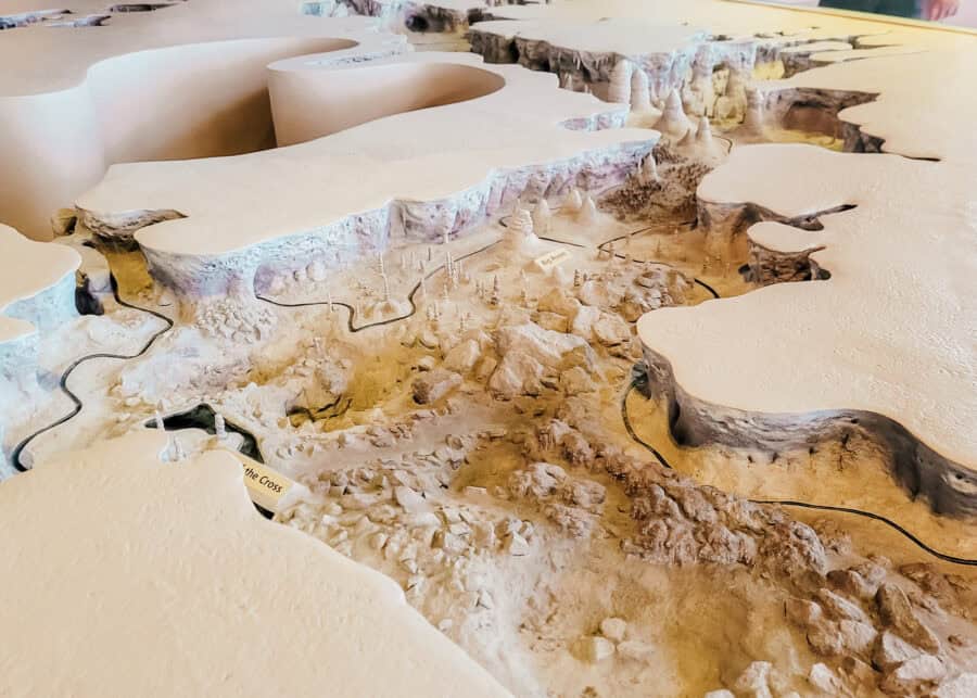 A detailed scale model of Carlsbad Caverns shows layered cave chambers with rounded ceilings and clusters of stalagmites below. Small labels read โBig Roomโ and โBat Cave Cross Section,โ and a winding path illustrates the layout through the formations.