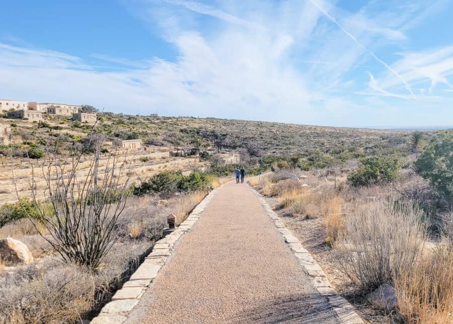 A paved walking path stretches through a dry desert landscape with shrubs and cacti on either side under a wide blue sky. Two people walk along the path toward low stone buildings and rolling hills in the distance.