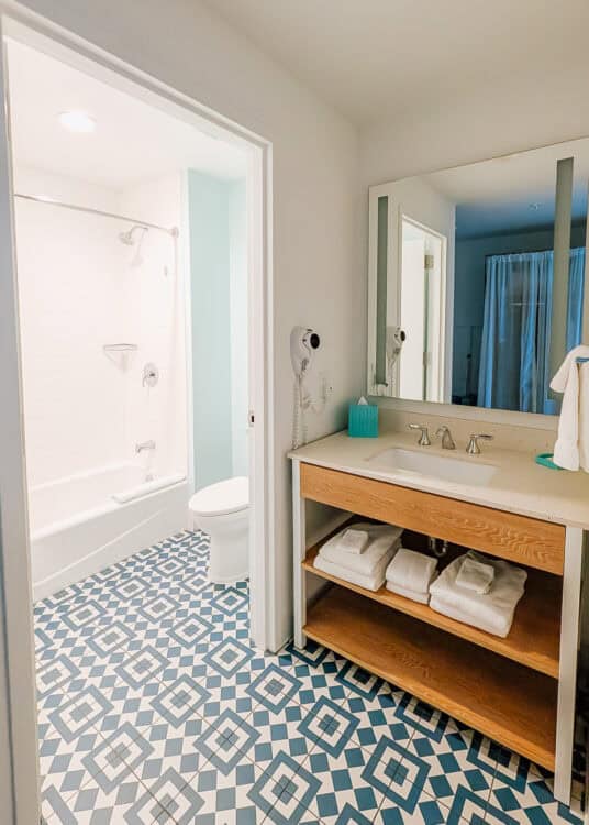 right hotel bathroom with a white sink and light wood vanity stocked with folded towels, next to a separate shower and bathtub area with blue and white patterned tile flooring. The clean coastal design and functional layout are highlighted in this endless summer surfside review.